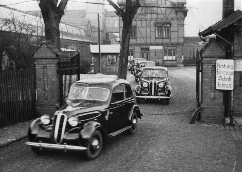 A black-and-white photograph of new BMW saloons driving out through the gate of the Eisenach car works.