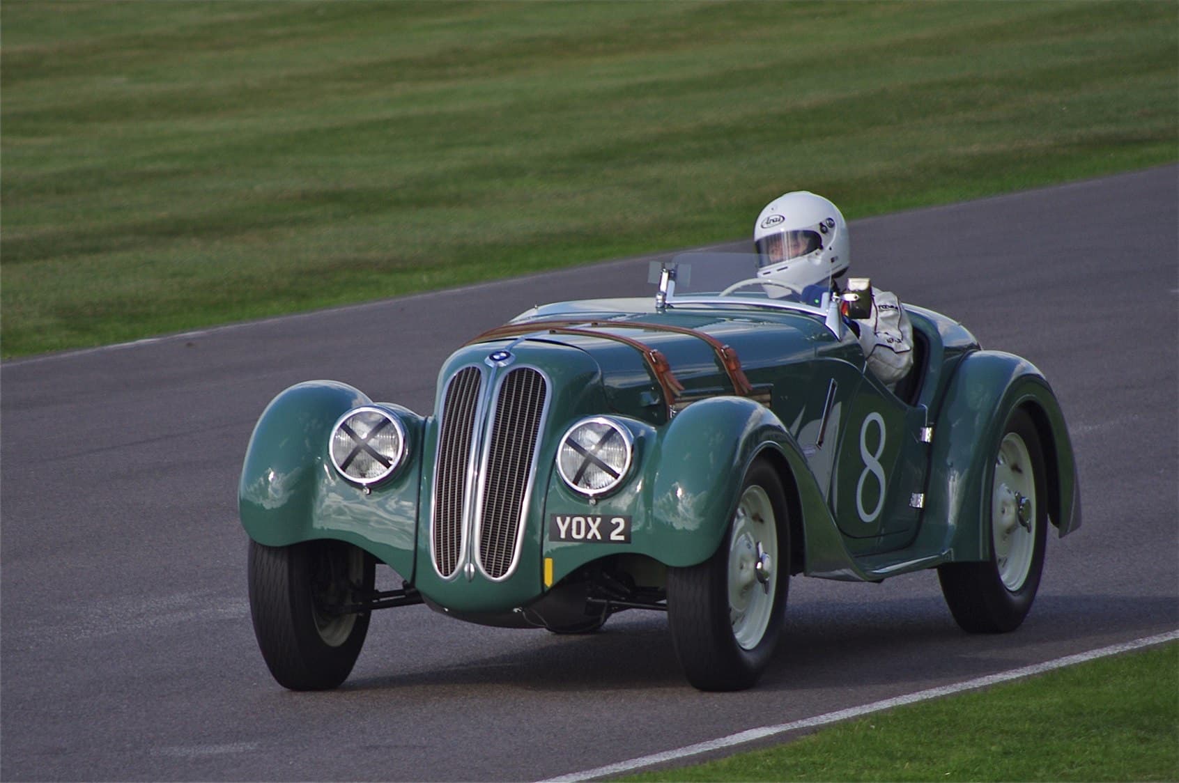 A green BMW 328 roadster with race number 8 cornering on a grass-edged circuit, its driver in a white helmet.