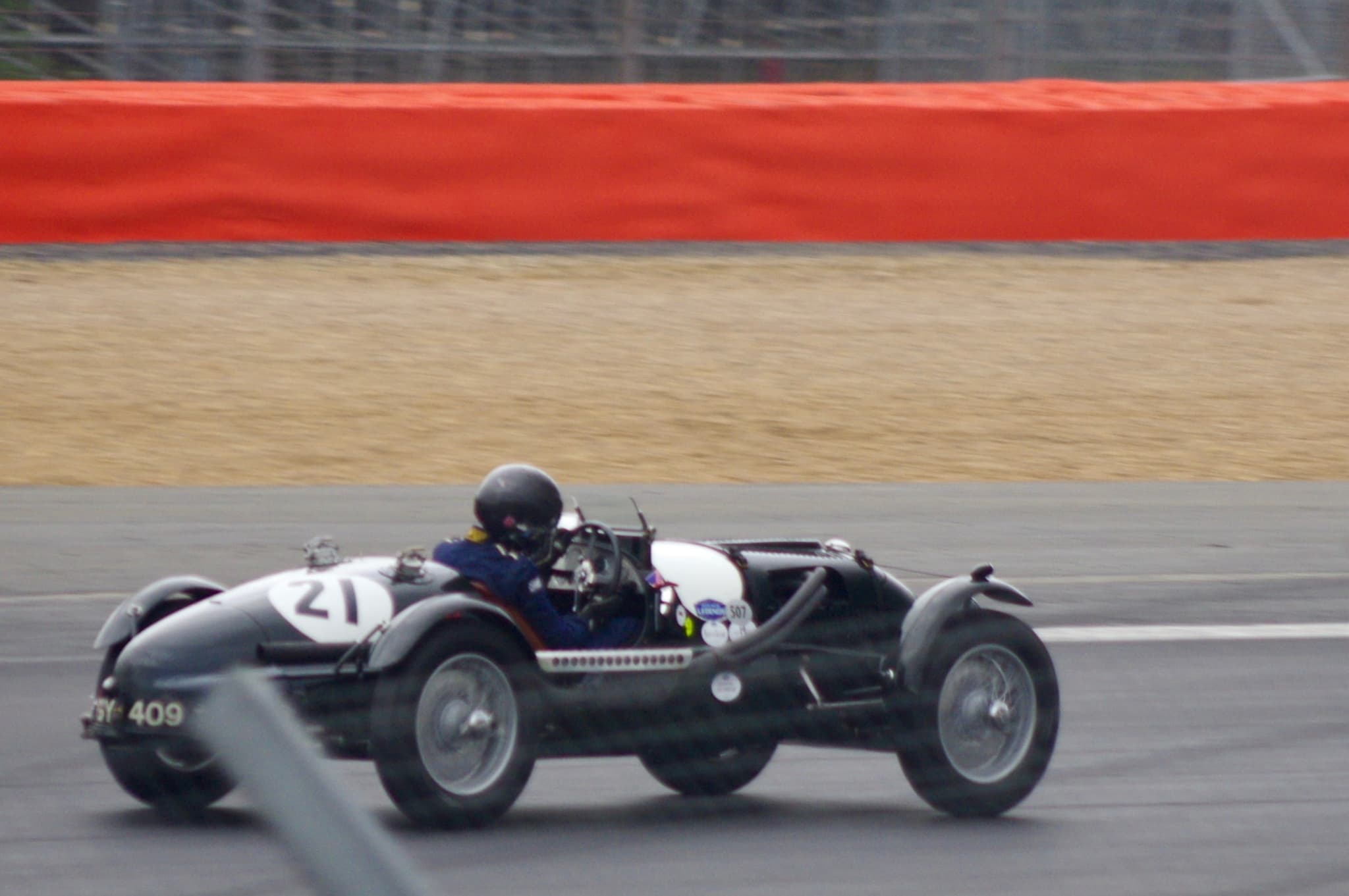 A dark Aston Martin pre-war sports car with race number 21 at speed on a circuit, a fence post blurred across the foreground.