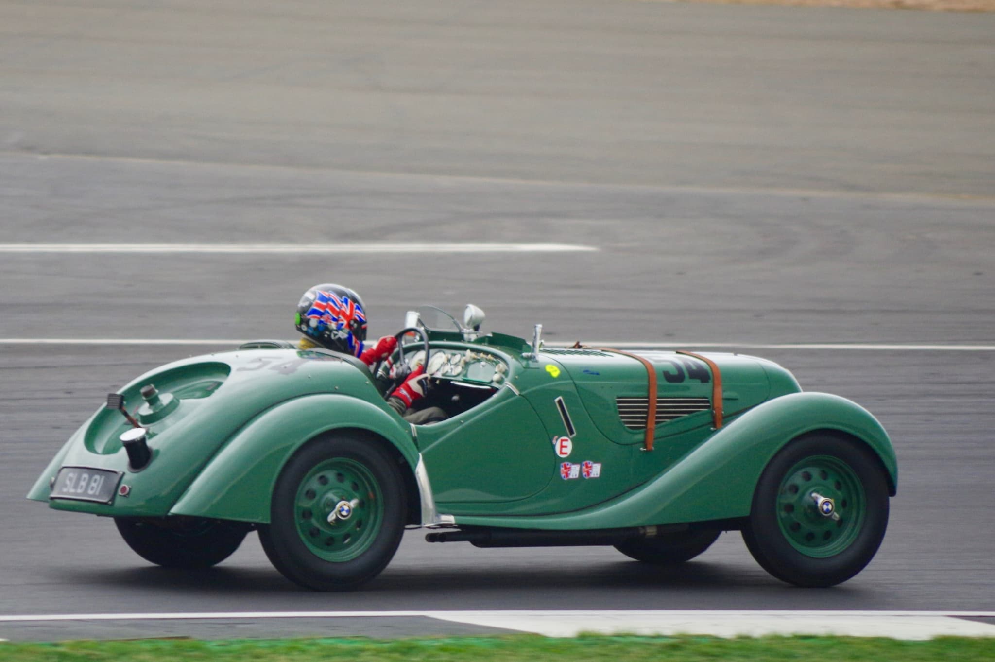 A green BMW 328 roadster numbered 54, seen from its right side at speed on a grey circuit, two leather straps over its bonnet and its driver in a helmet painted with the Union flag.