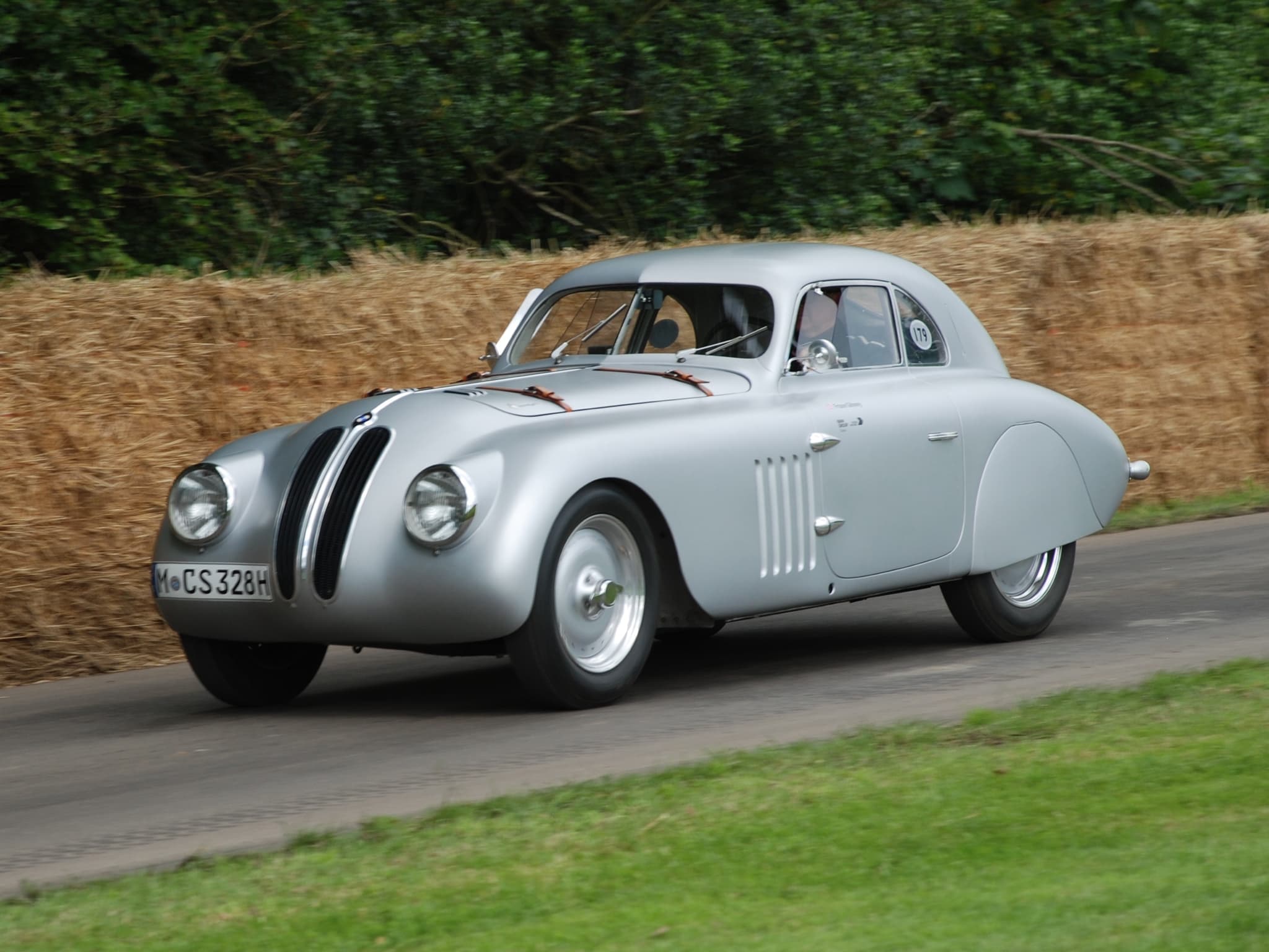 A silver BMW 328 fastback racing coupé driving uphill past straw bales and a hedge.