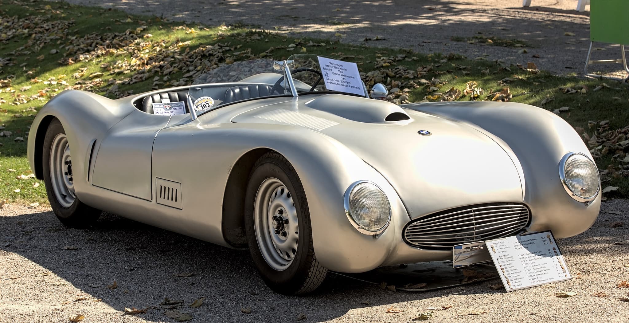 A silver streamlined Werkmeister racing car on a gravel drive under trees, with an information board at its nose.