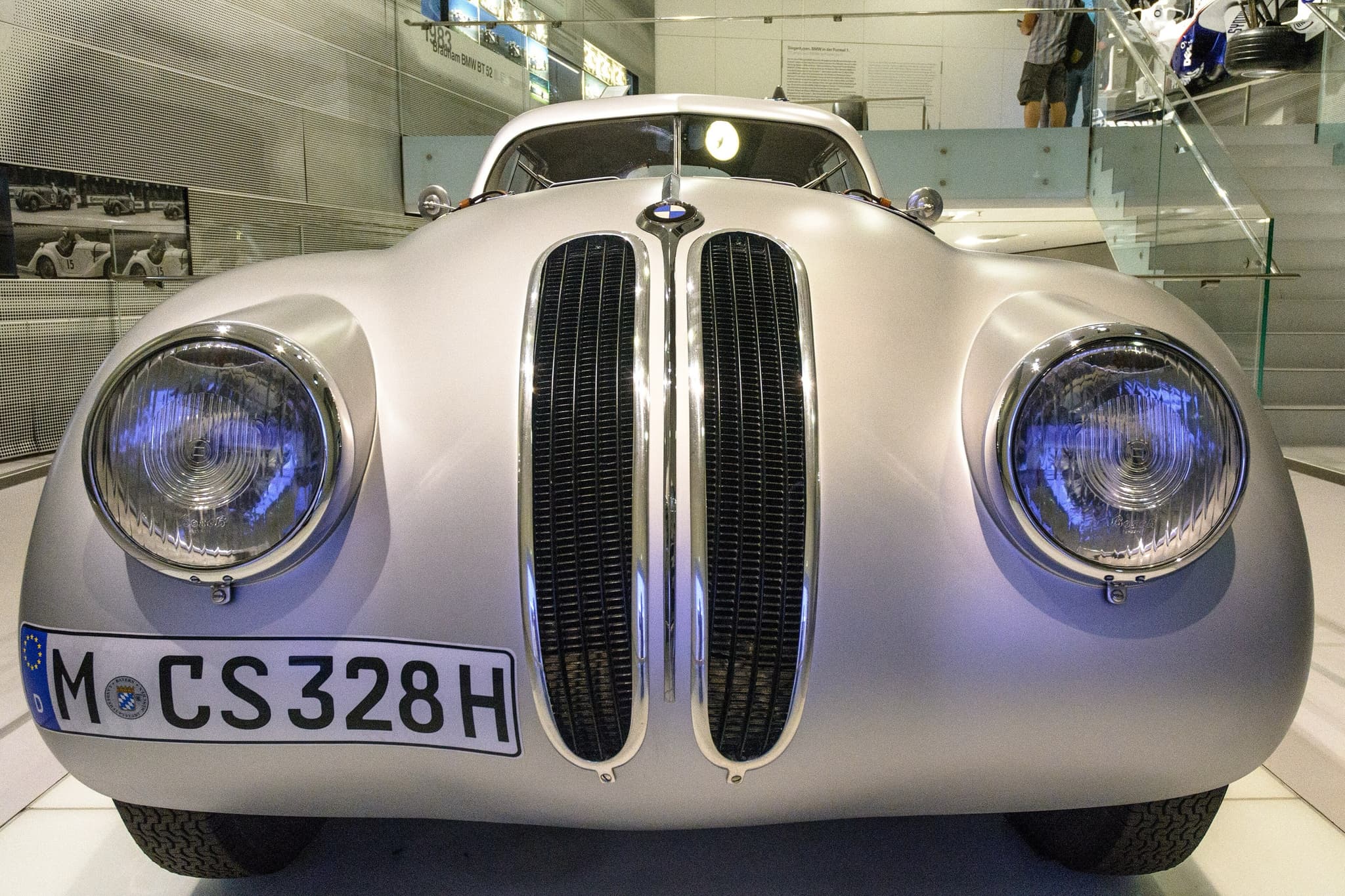 A silver BMW 328 racing coupé with a fastback roof seen nearly head-on in a museum, leather straps across its bonnet.