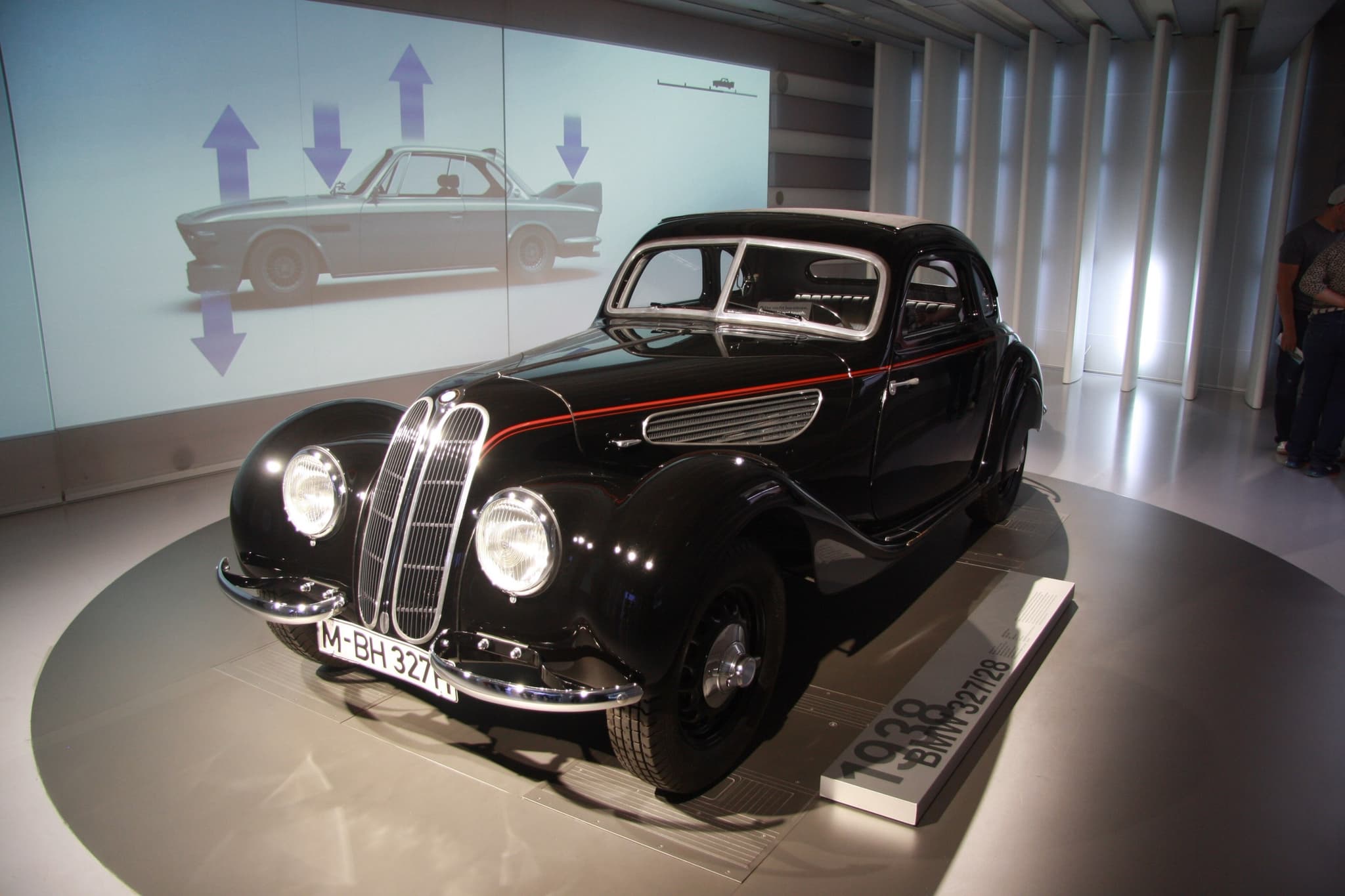 A black BMW 327/28 coupé with a red side line on a museum turntable beside a plinth reading 1938 BMW 327/28.