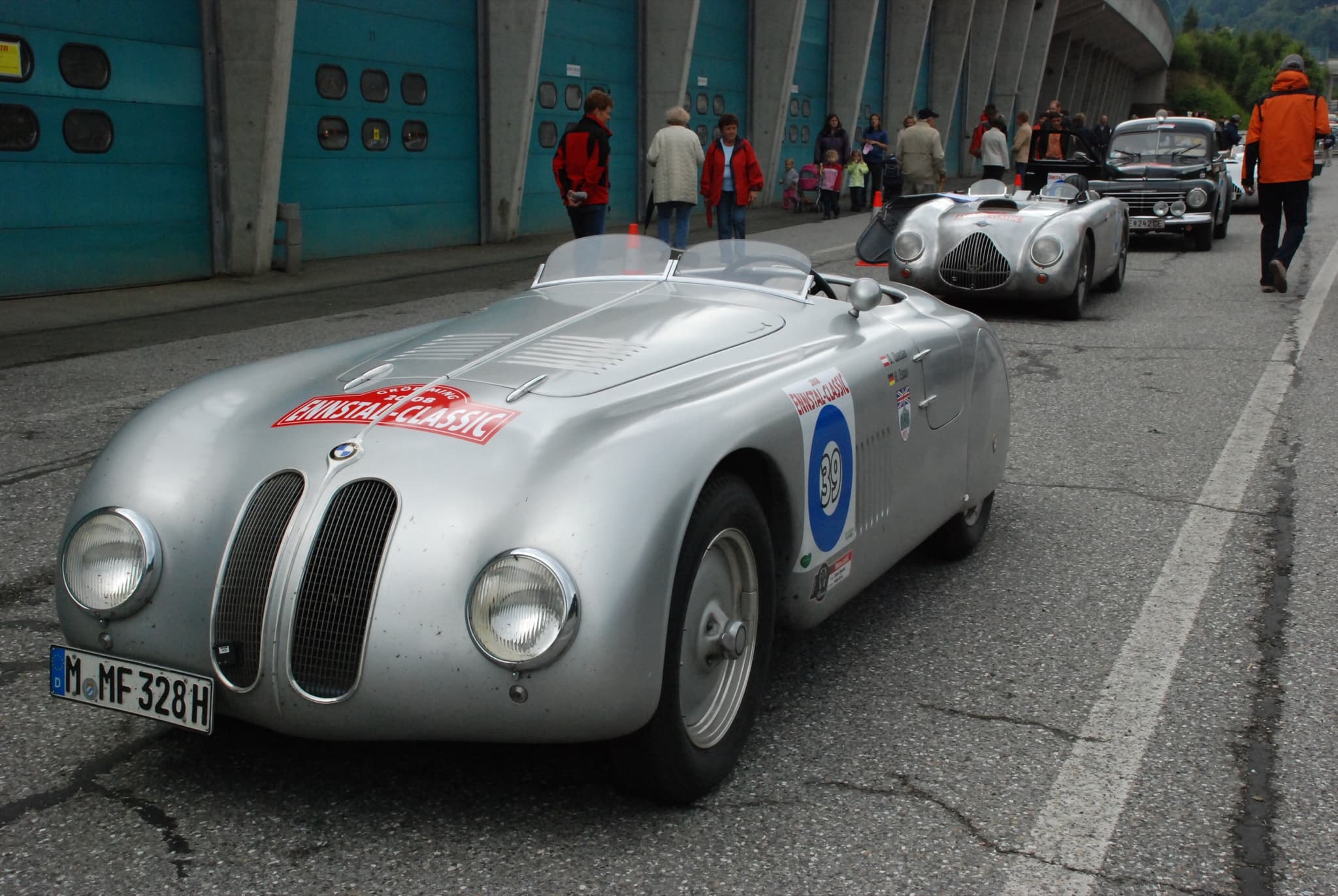 A silver streamlined BMW roadster with an event plate on its bonnet in a pit lane, other classic cars behind.