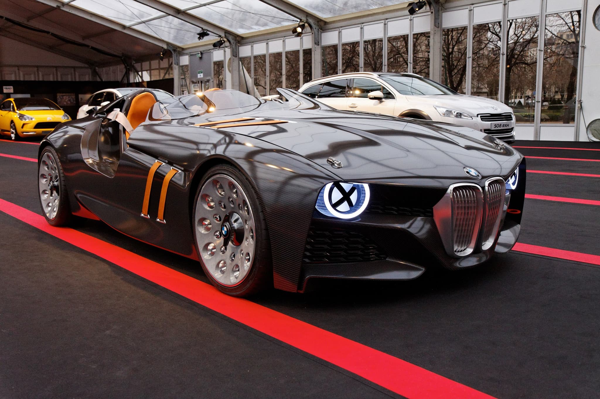 The BMW 328 Hommage concept car from the right front in an exhibition tent, its X-lit headlamp and tan bonnet straps glowing. Opens a larger view