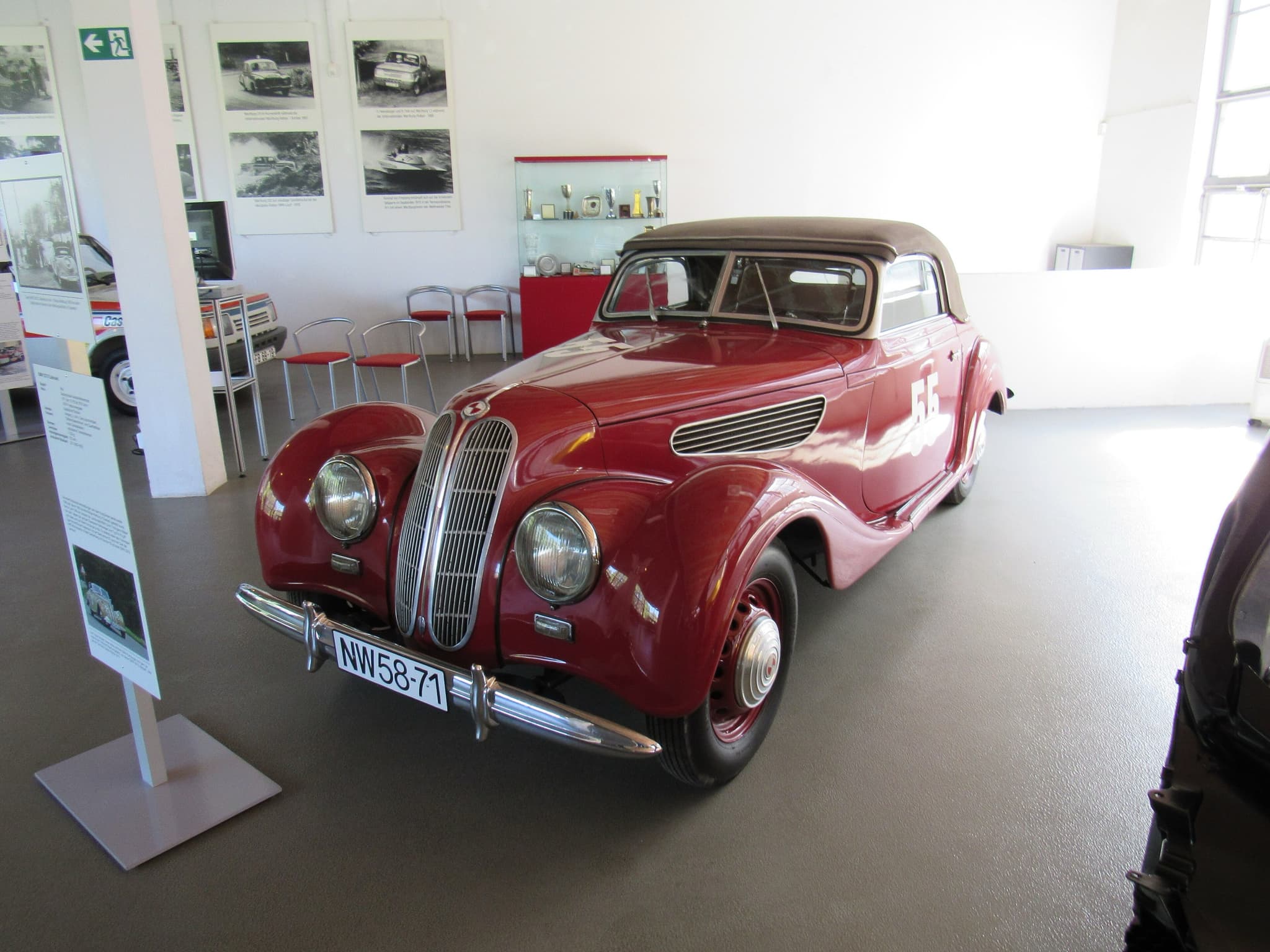 A red EMW 327 cabriolet with a brown hood in a museum, a trophy cabinet and enlarged rally photographs behind.