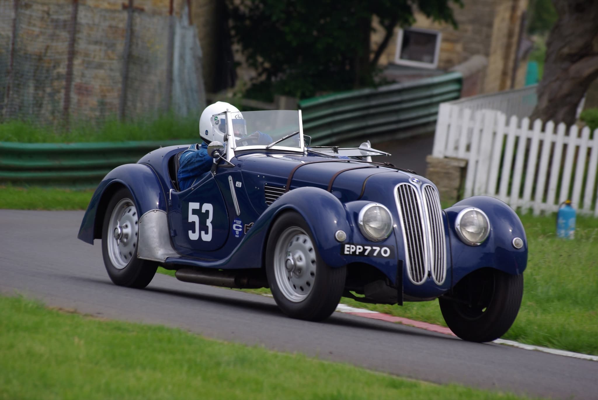 A dark-blue BMW 328 roadster with the number 53 on its door, climbing a hill-climb course between a stone wall and a white picket fence, its driver in a white helmet.