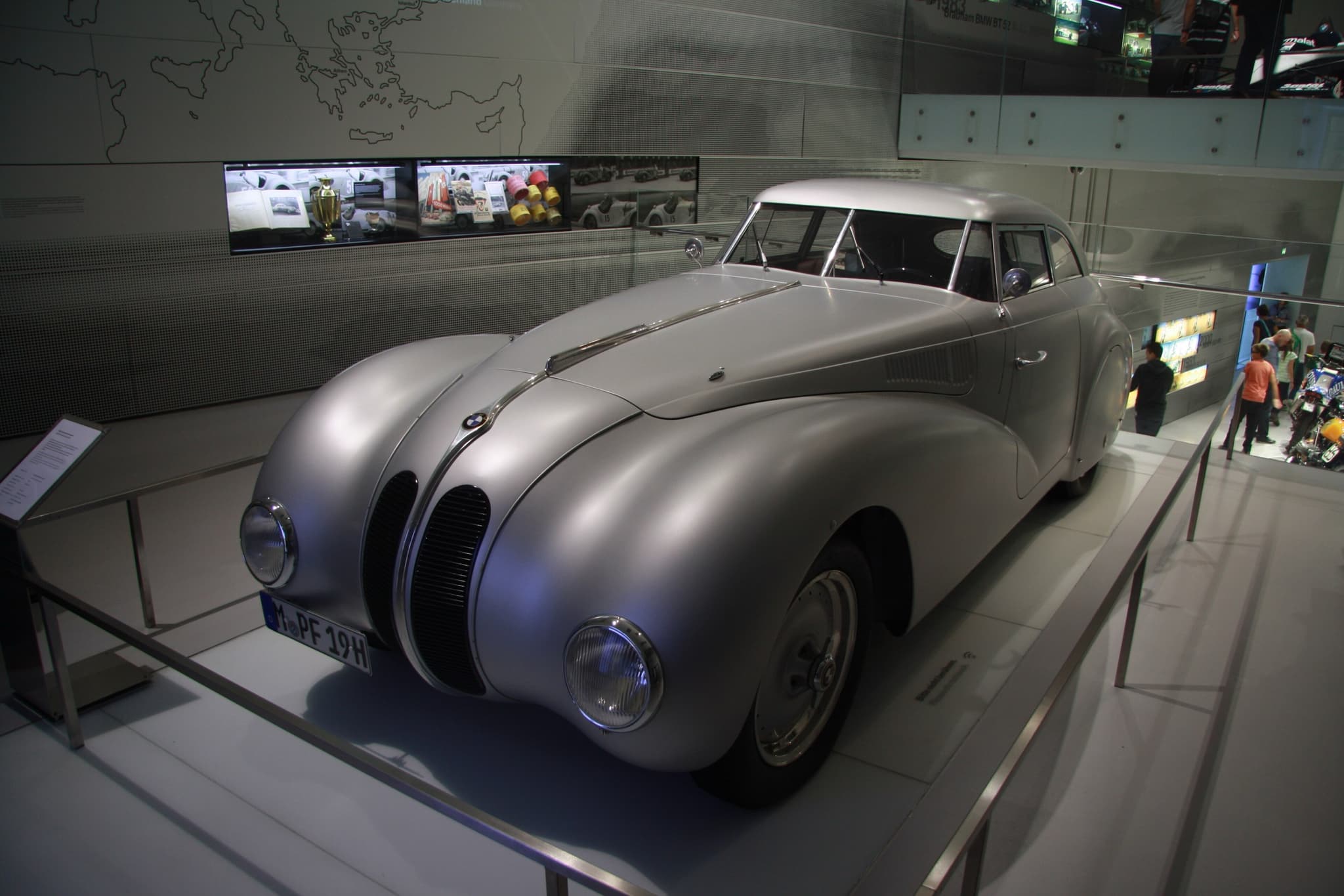 A silver streamlined BMW coupé on a museum plinth behind a steel rail, seen from the front left, with full-width front wings, twin kidney grilles and a long fastback roof.