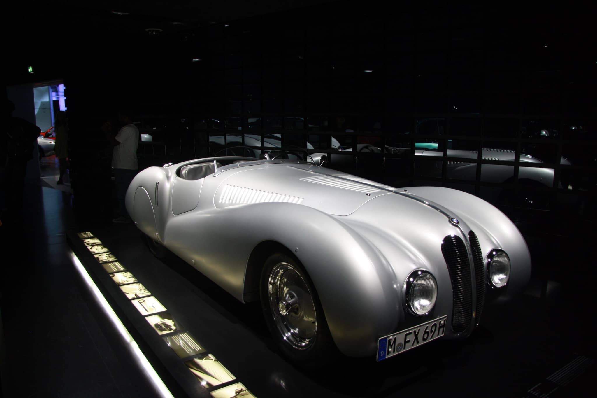 A silver roadster with a louvred bonnet and faired wings, under spotlights in a darkened museum hall.
