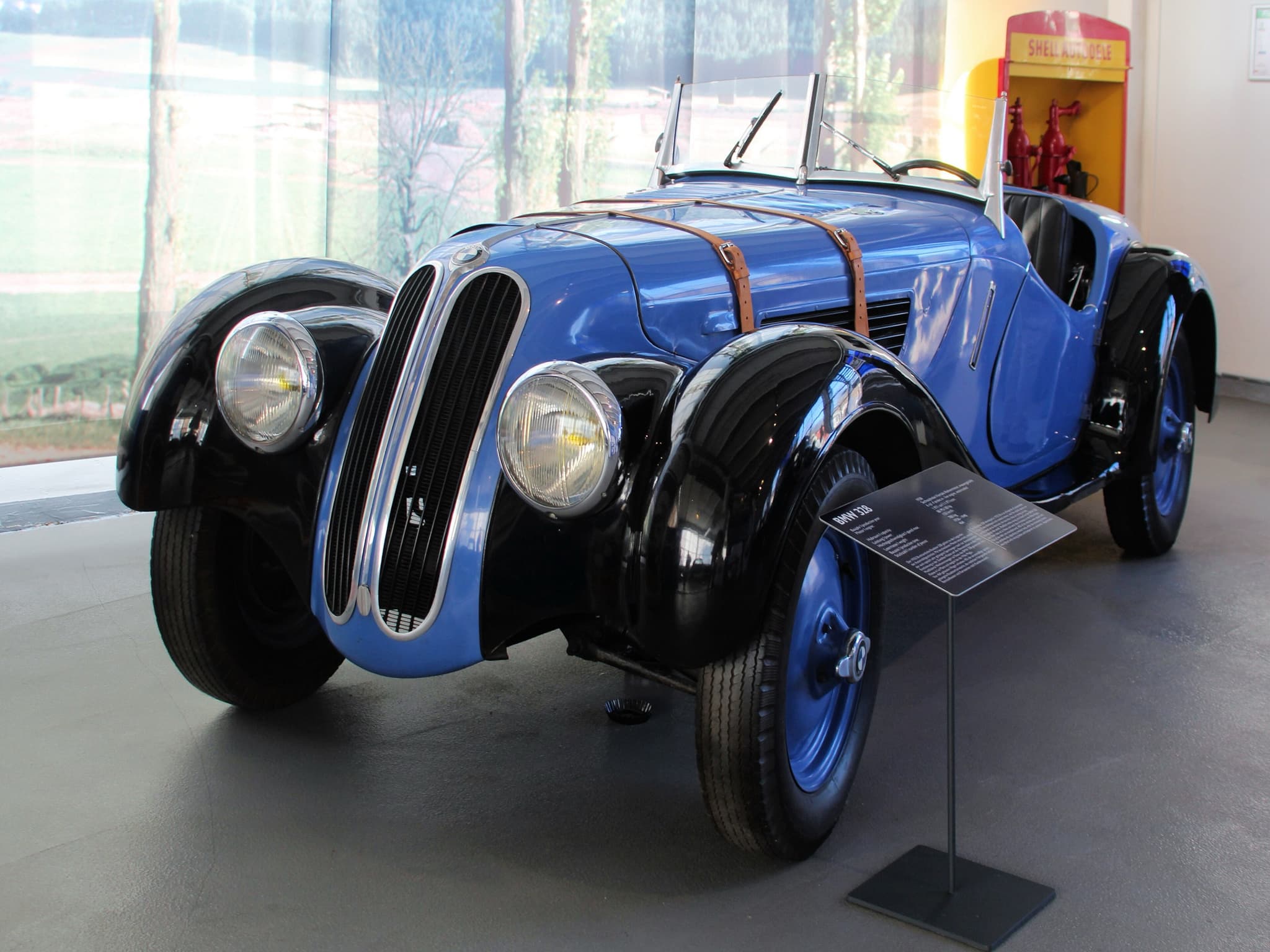 A blue and black BMW 328 roadster seen from the front left on a grey museum floor, with its twin kidney grilles, headlamps set into black wings and two leather bonnet straps, a small information stand in front of it.