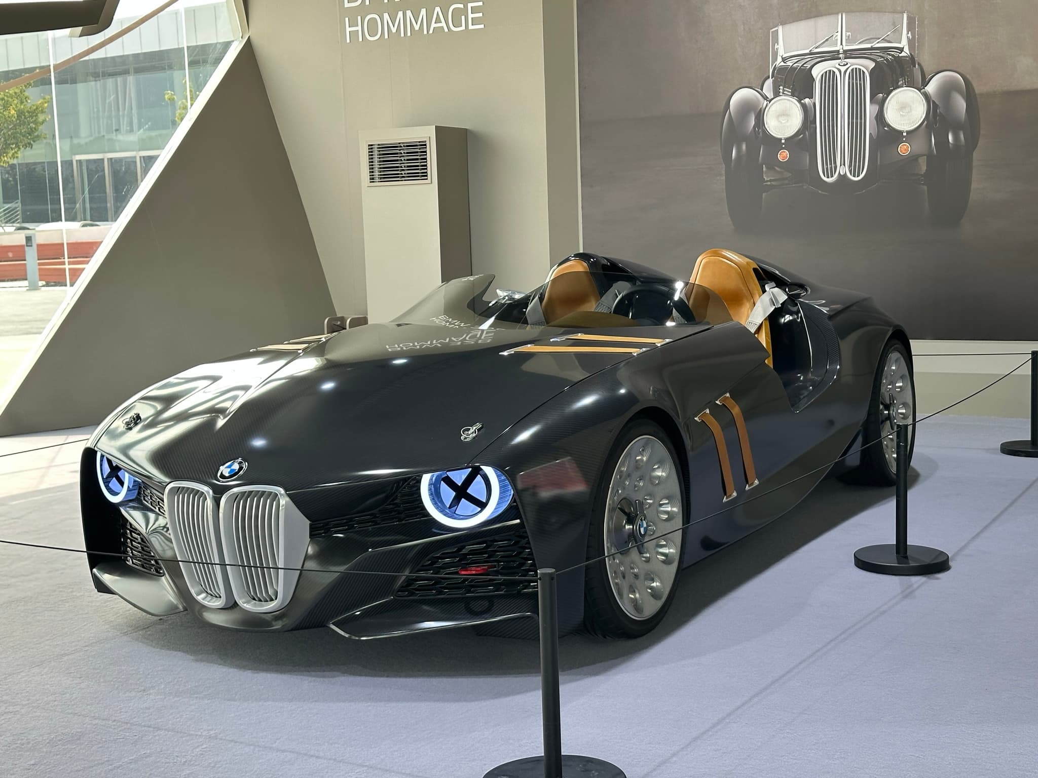 The BMW 328 Hommage concept car behind a rope at an exhibition, a large photograph of a 1930s BMW 328 on the wall behind.