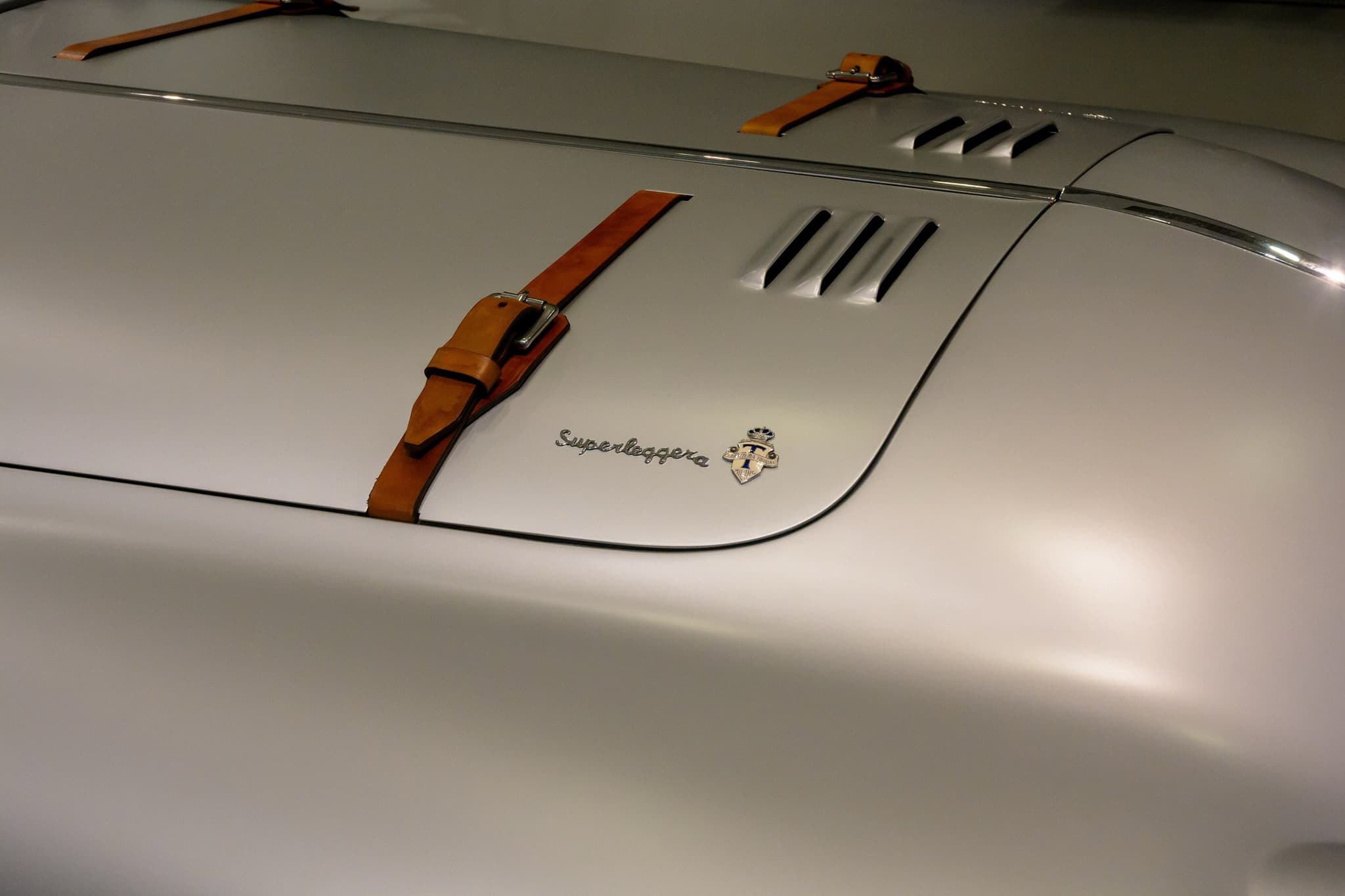 Close view of a silver bonnet with a chrome 'Superleggera' script and Touring badge, a leather strap and louvres.