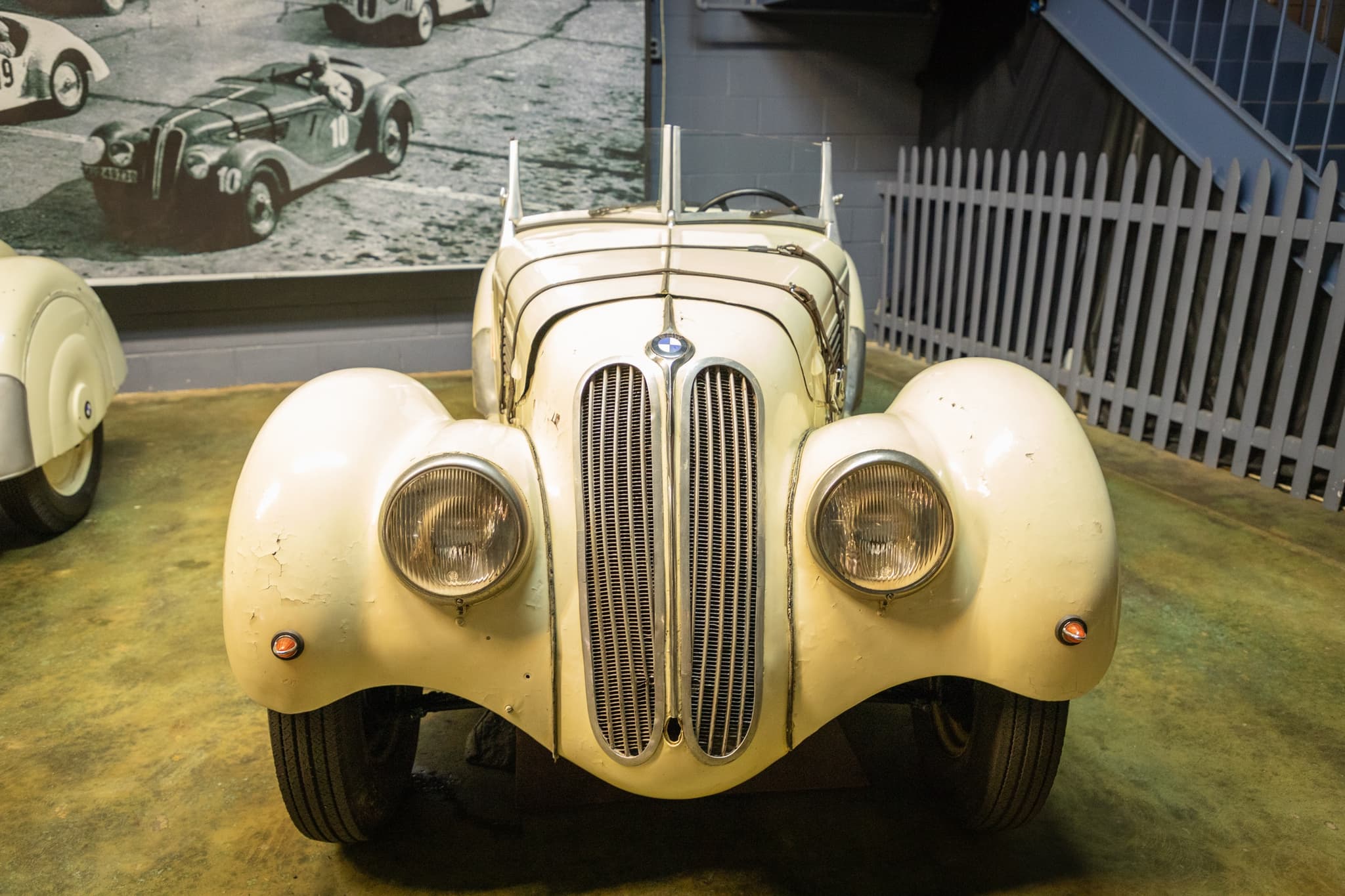 An unrestored ivory BMW 328 roadster seen head-on in a museum, its paint crazed, a blue staircase and a white fence behind.