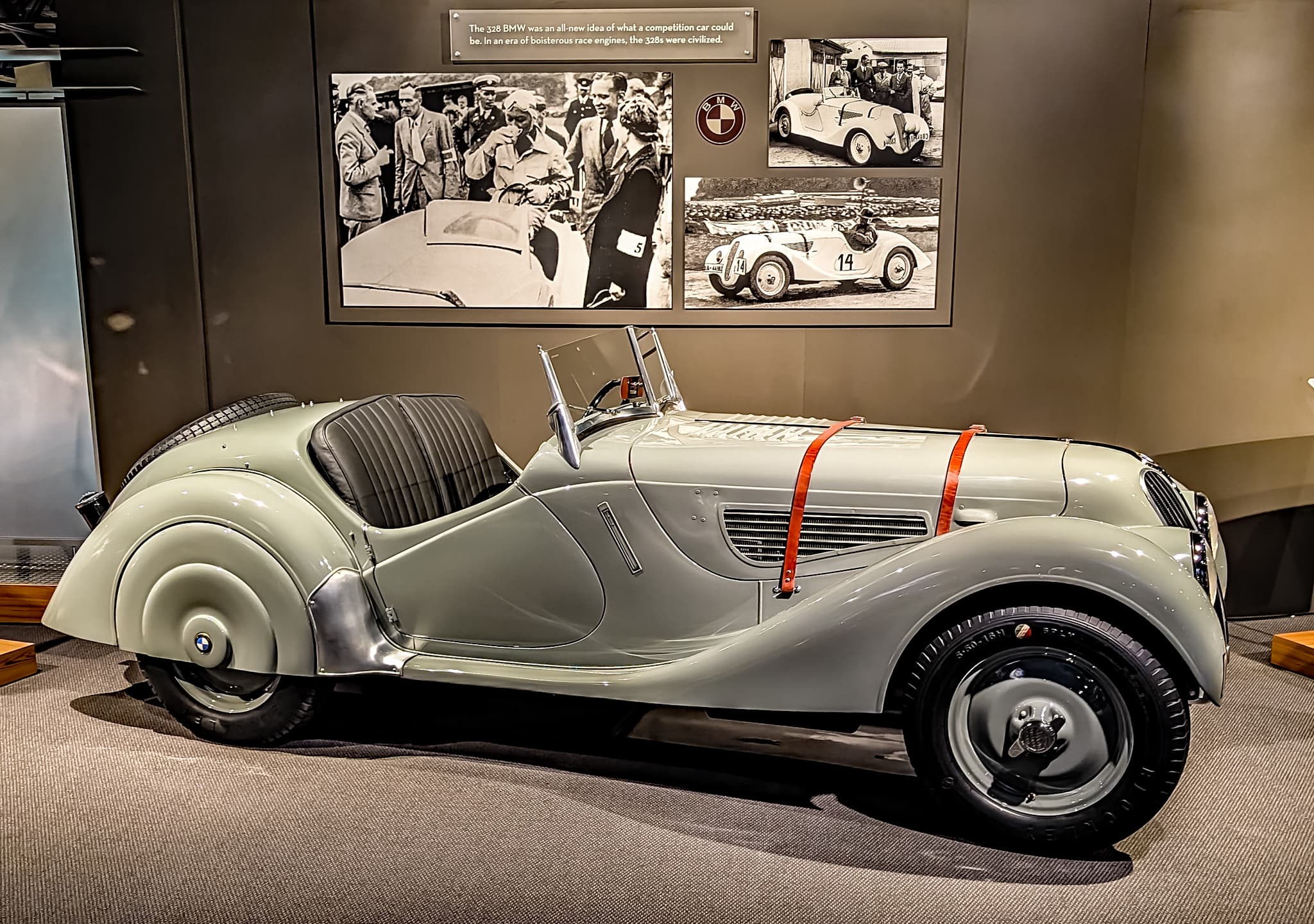 A grey BMW 328 roadster in profile in a museum gallery, enlarged period race photographs on the wall above it.