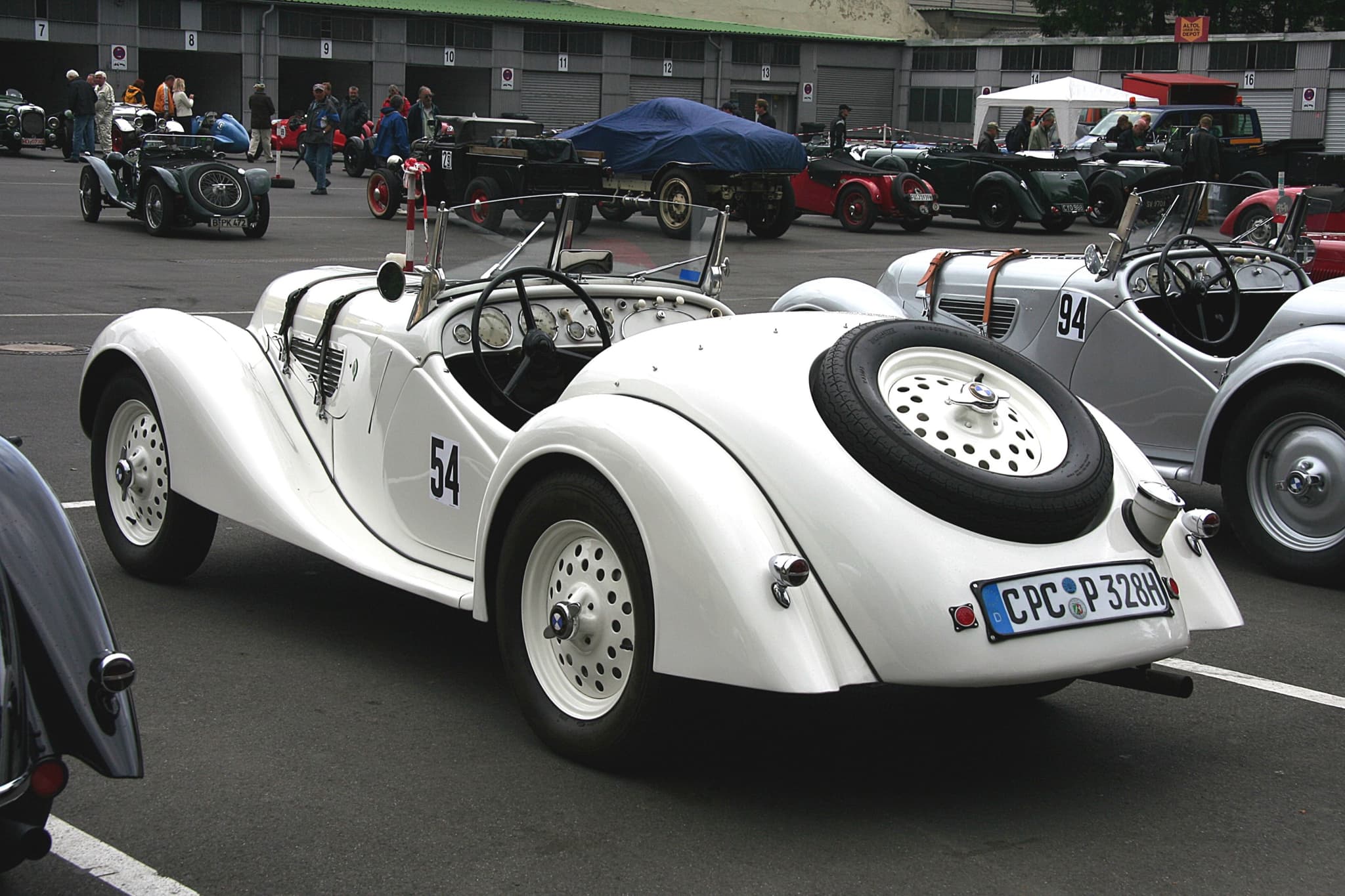 A white BMW 328 roadster with race number 54 from the left rear in an old racing paddock, beside a silver 328 and other pre-war cars. Opens a larger view