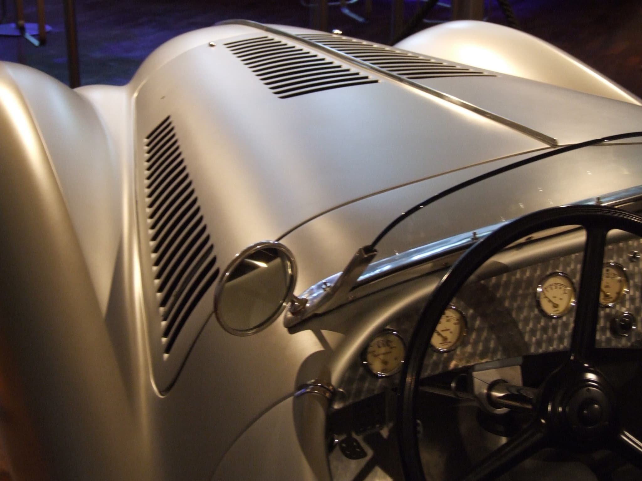 Close view of a silver streamlined roadster's louvred bonnet, mirror and cockpit with an engine-turned dashboard, the wheel on the right.
