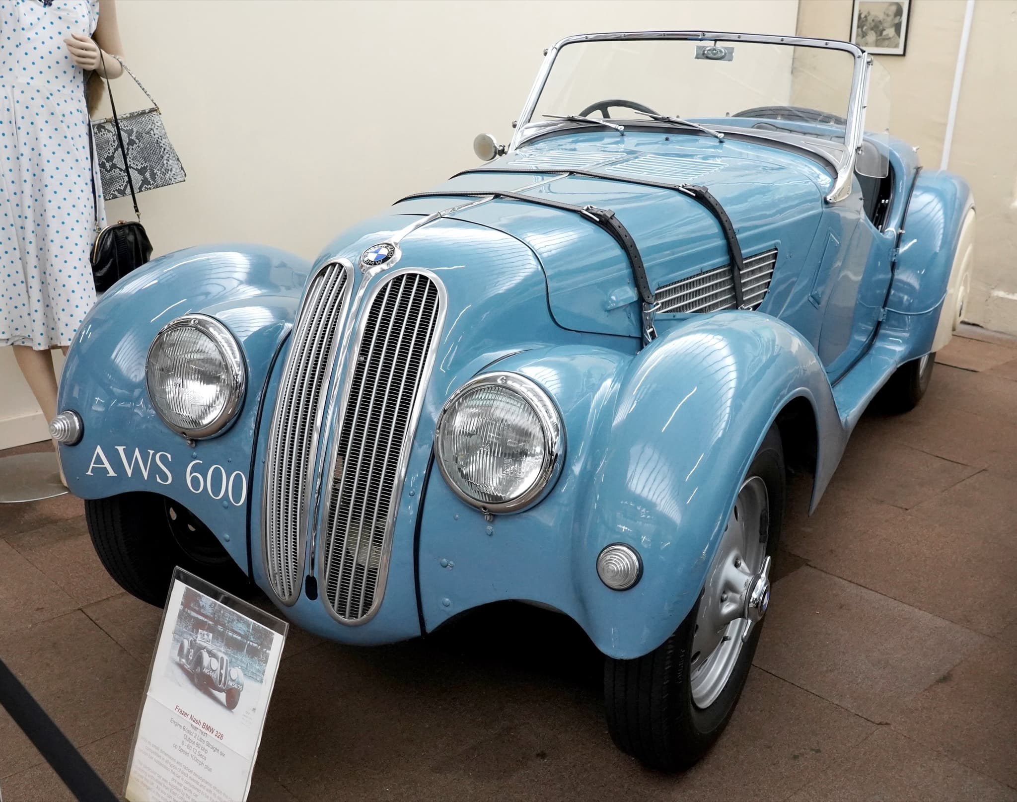A light-blue BMW 328 roadster in a small museum with a card reading 'Frazer Nash BMW 328' before it and a dressed mannequin beside it.