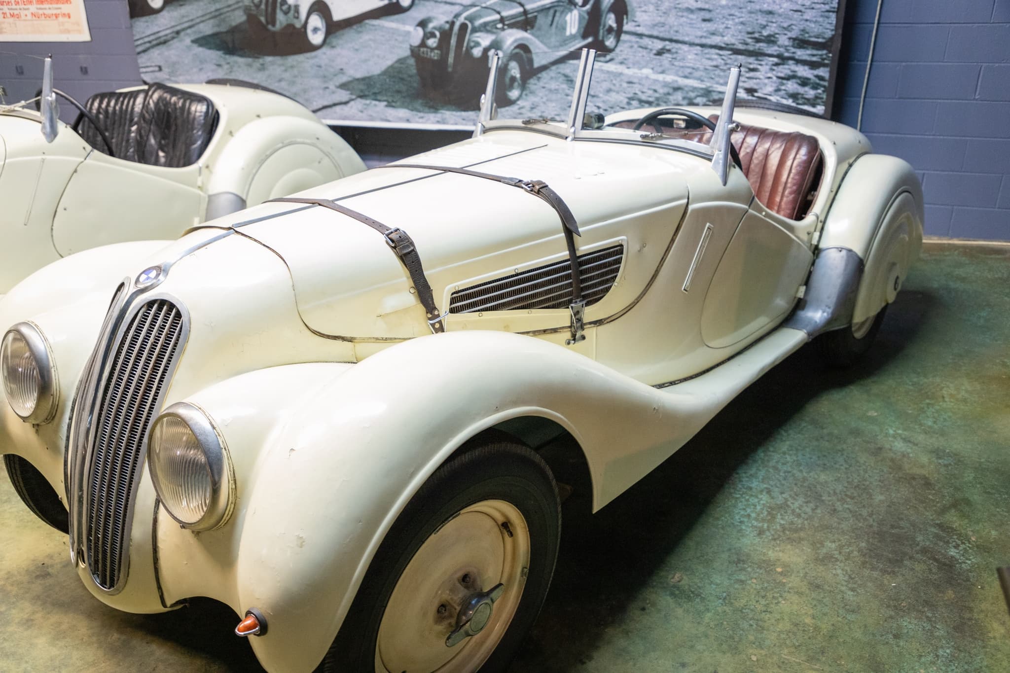 An ivory BMW 328 roadster with worn, crazed paint and red seats in a museum diorama, a mural of a 1930s race grid behind it.