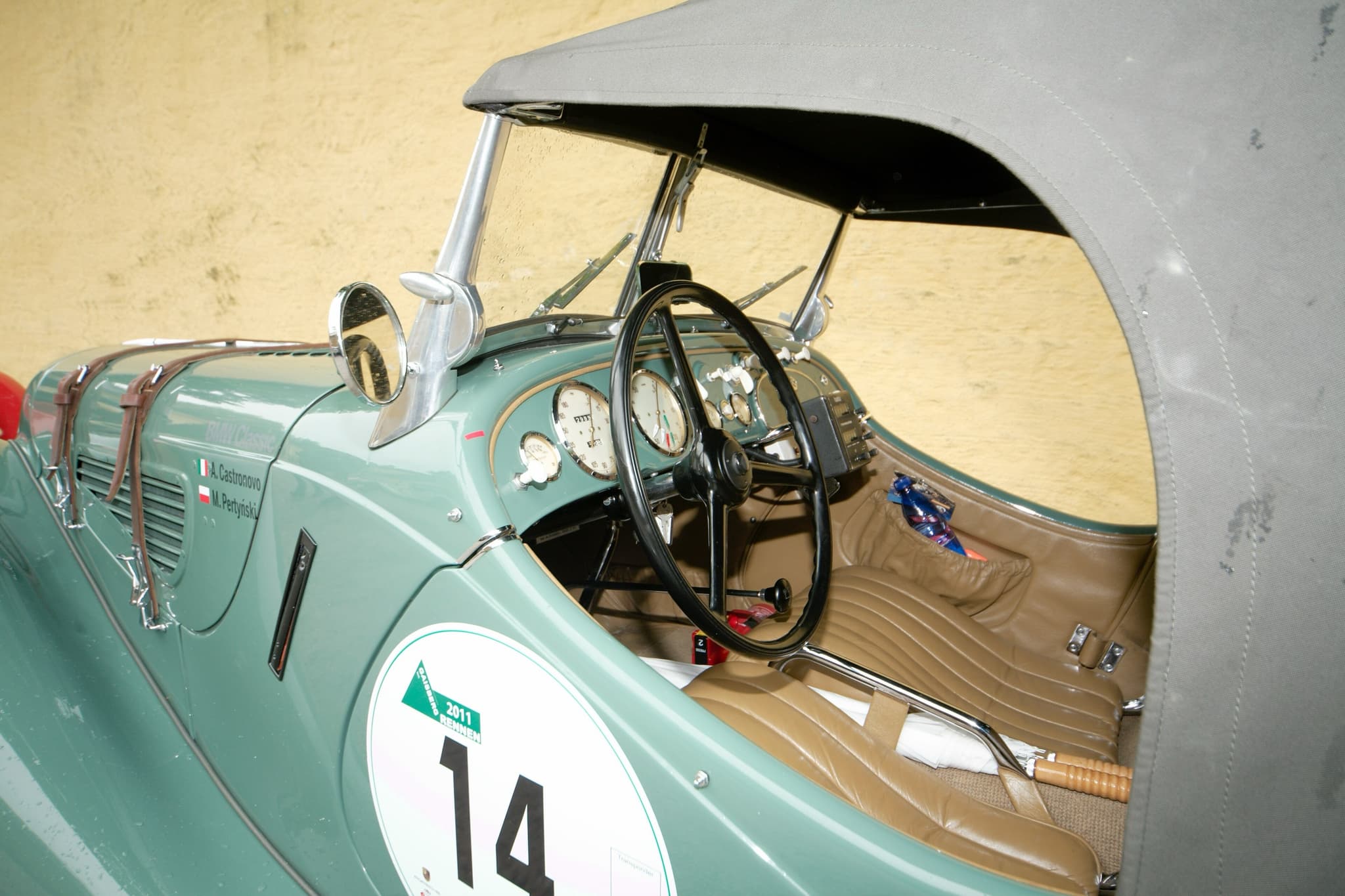 The cockpit of a green BMW 328 under its hood, with a three-spoke wheel, dials, tan leather seats and a rally trip computer.