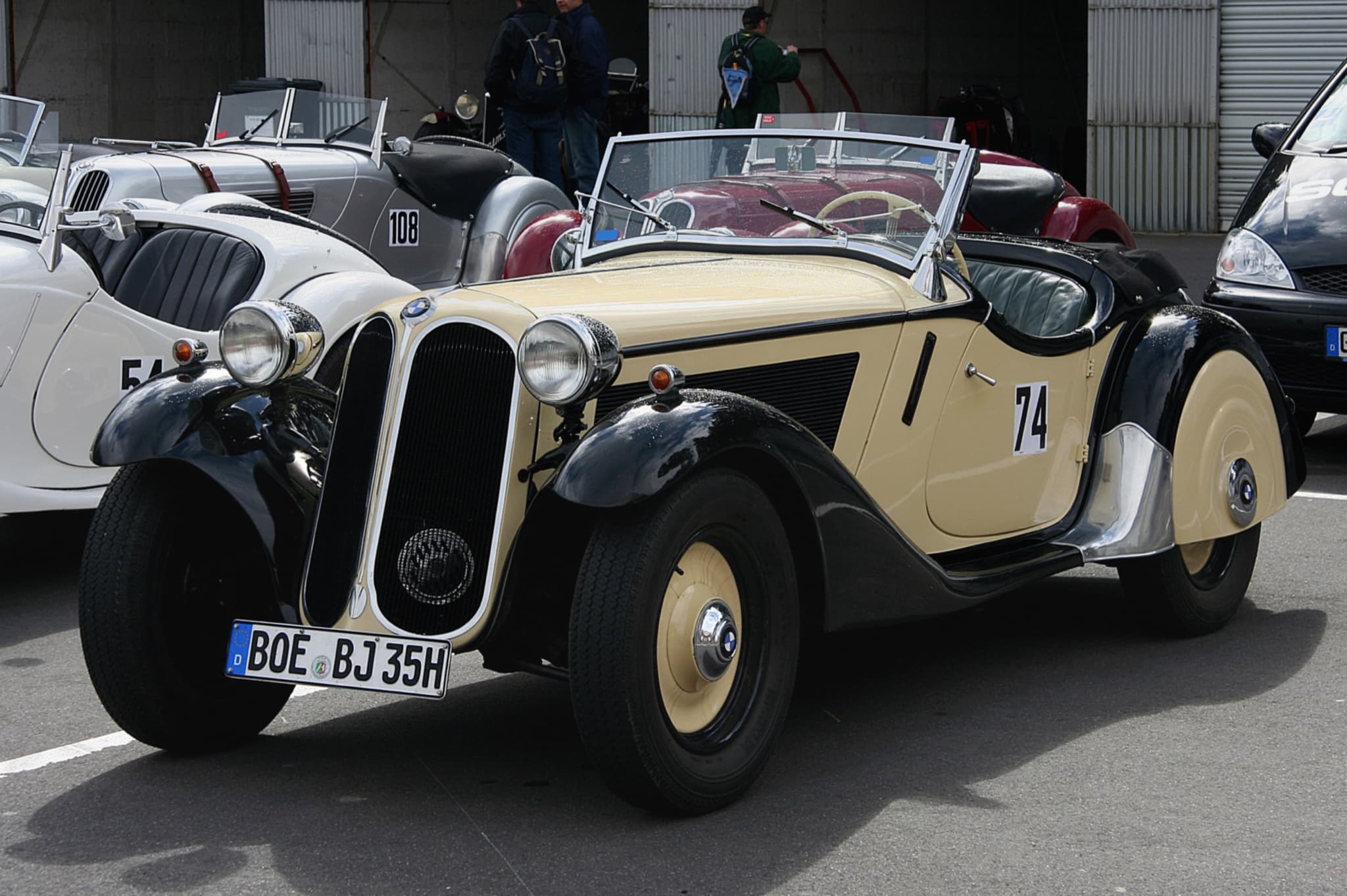A cream-and-black BMW 315/1 roadster with race number 74 in a racing paddock, two BMW 328s beside it.