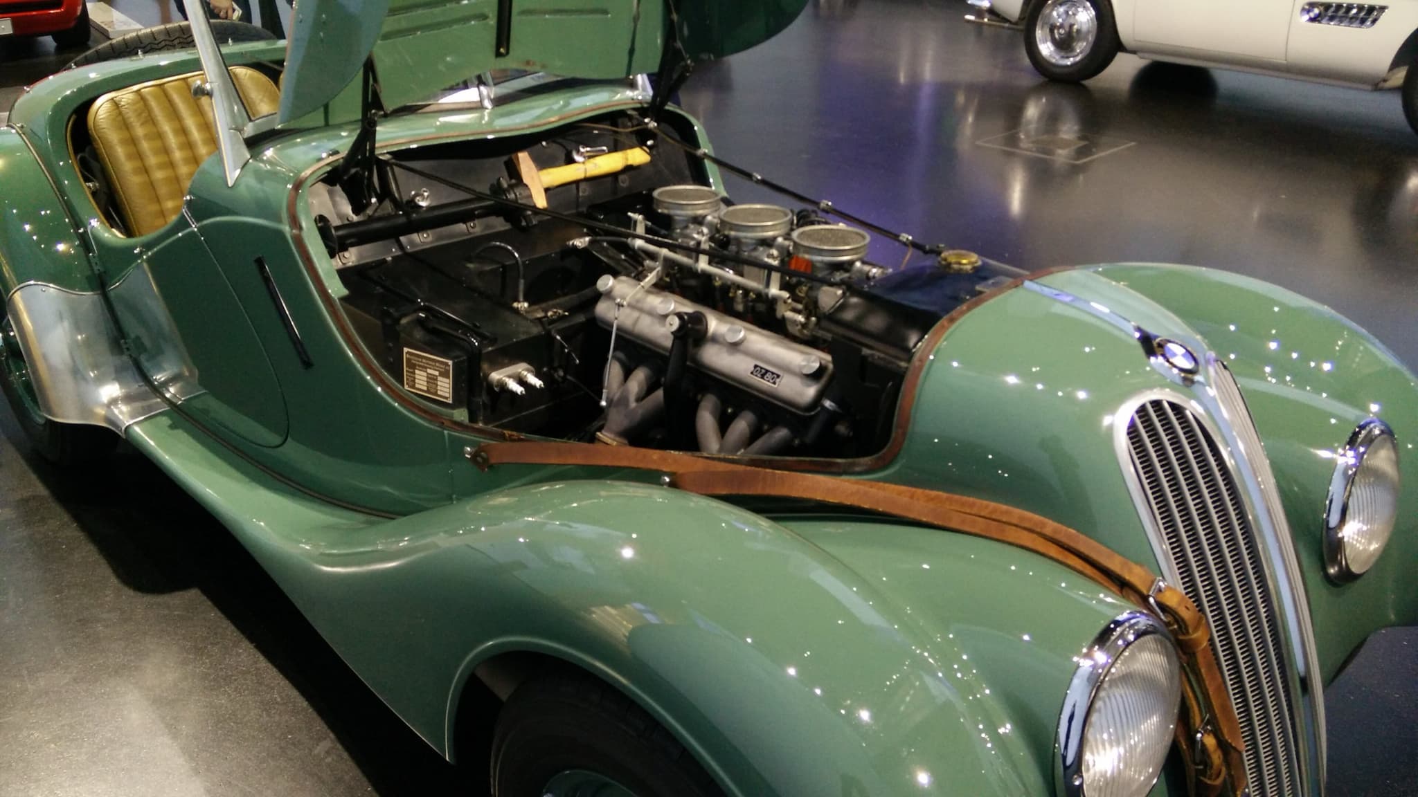 A green BMW 328 roadster with the side of its bonnet raised, showing the six-cylinder engine, its carburettors and the exhaust manifold.