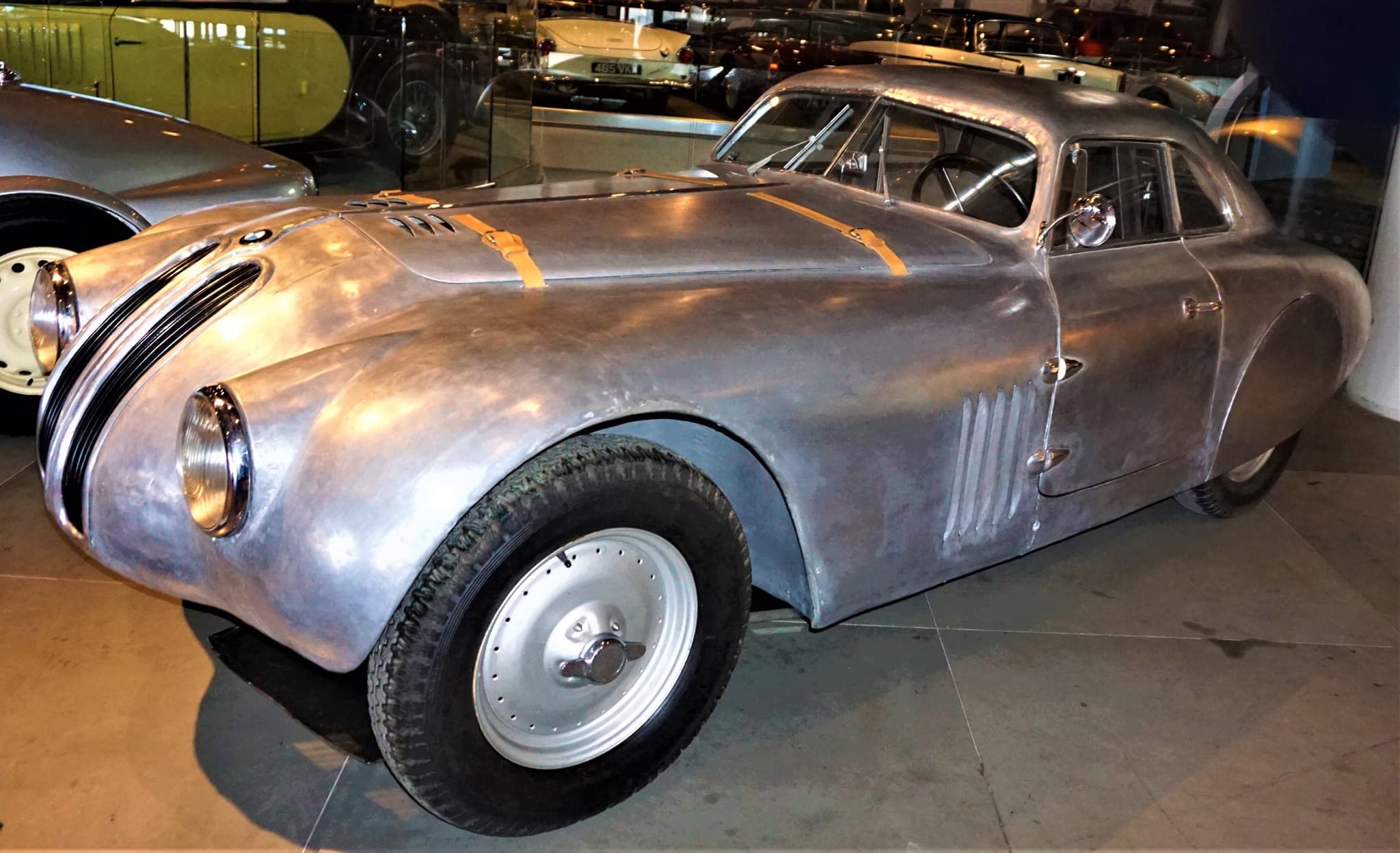A bare-aluminium closed coupé body on a BMW 328 base in a museum, with BMW kidney grilles and leather bonnet straps.