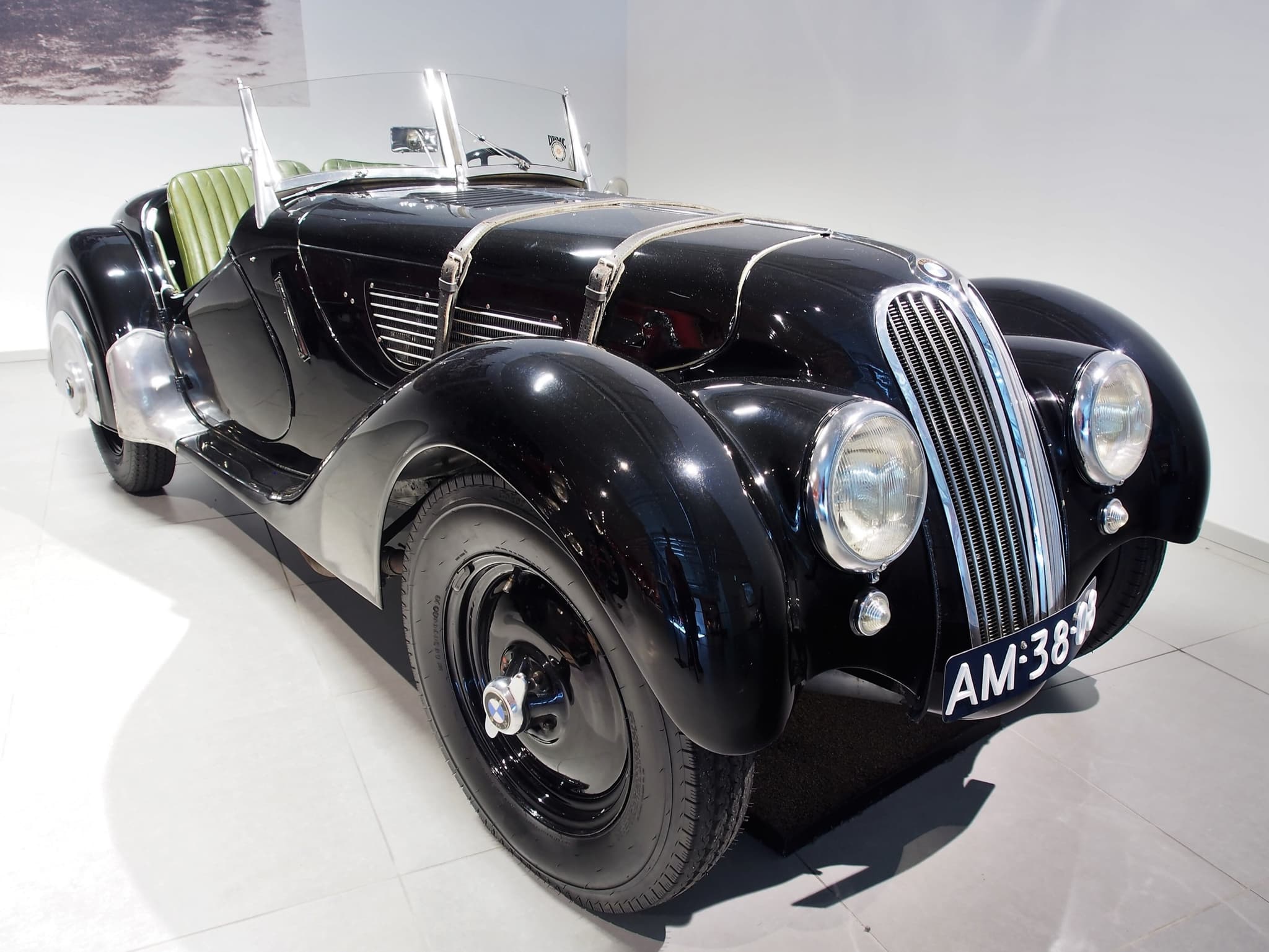 A black BMW 328 roadster with green leather seats in a white museum hall, seen low from the front right.