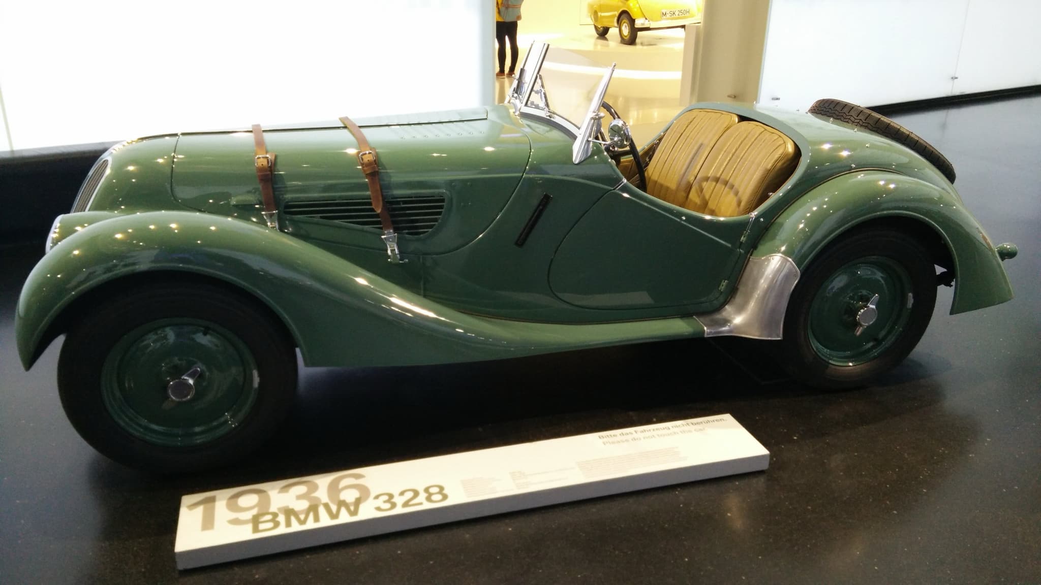 A green BMW 328 roadster in profile on a dark museum floor, with leather bonnet straps, louvres and tan seats.