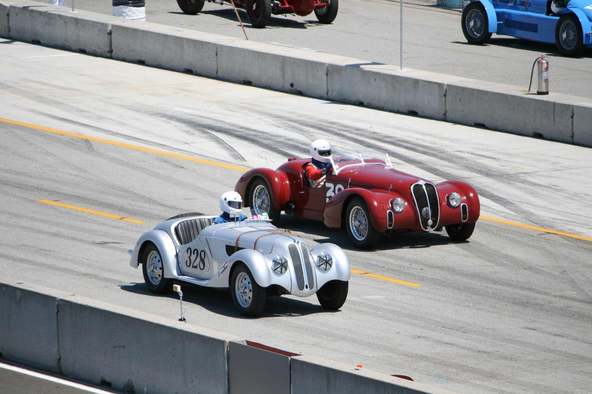 A silver BMW 328 roadster and a red Alfa Romeo sports car side by side in a pit lane, their drivers in white helmets.