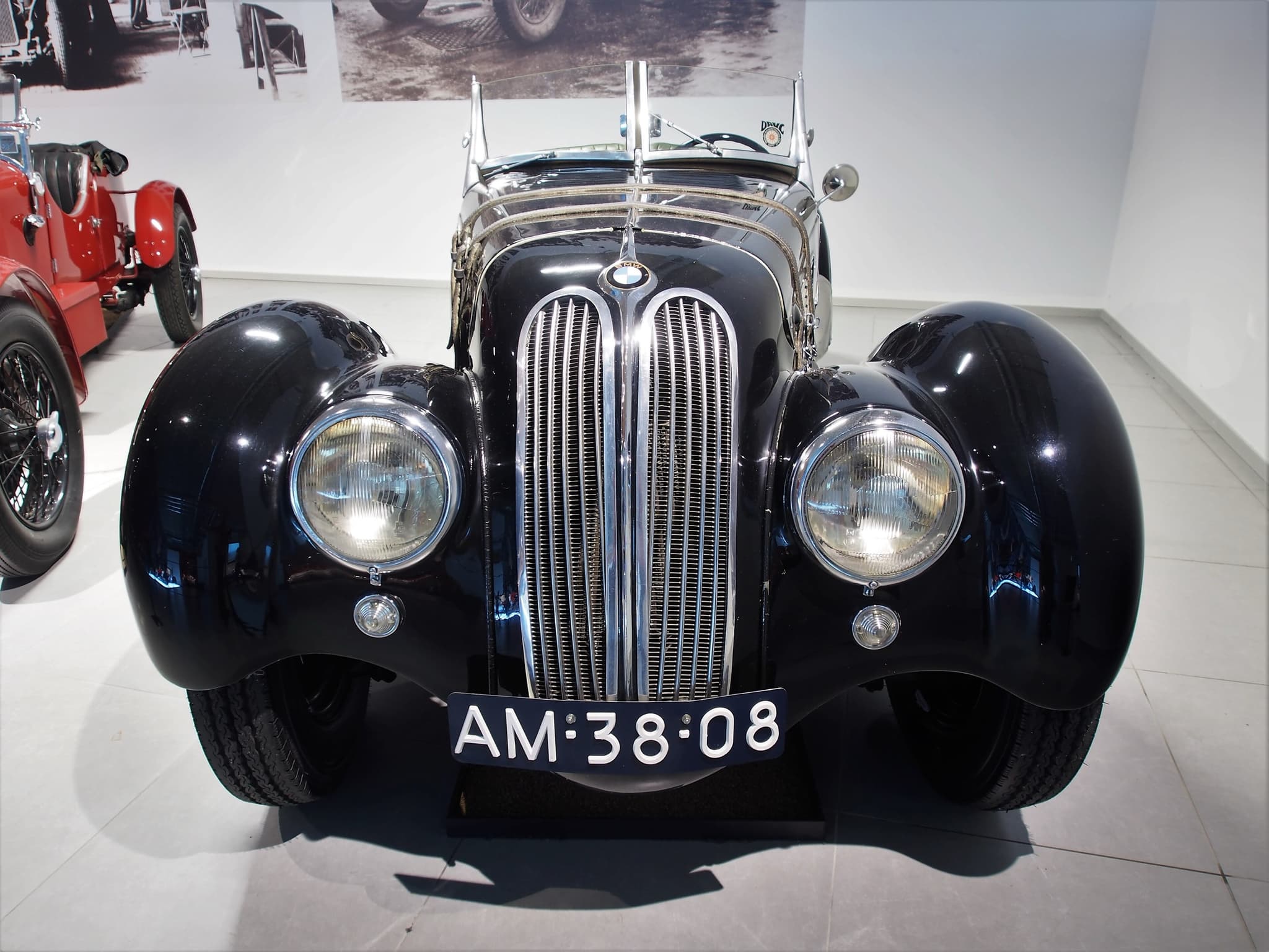 A black BMW 328 roadster seen square-on in a museum, its two tall narrow chrome grilles between round headlamps faired into the wings, a red vintage car cut by the left edge and enlarged period photographs on the white wall behind.