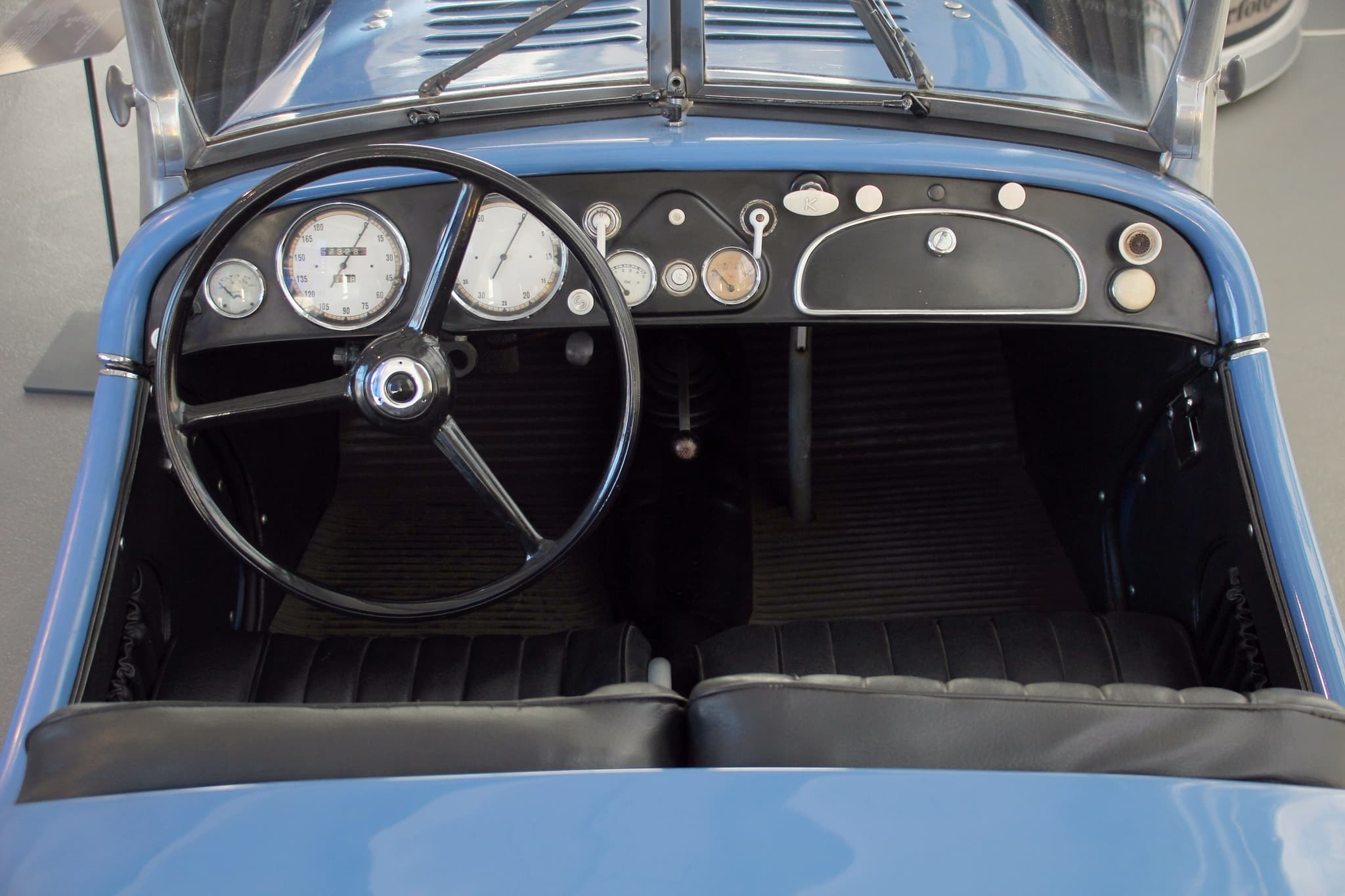 The cockpit of a BMW 328 from above: a black three-spoke steering wheel on the left, two large dials, three small gauges and a lidded glove box.
