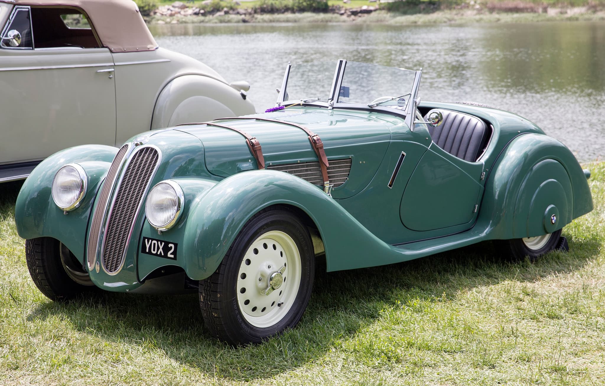A green BMW 328 roadster on grass by a river, showing its tall twin-kidney grille, headlamps faired into the wings, two leather bonnet straps and a vee windscreen.