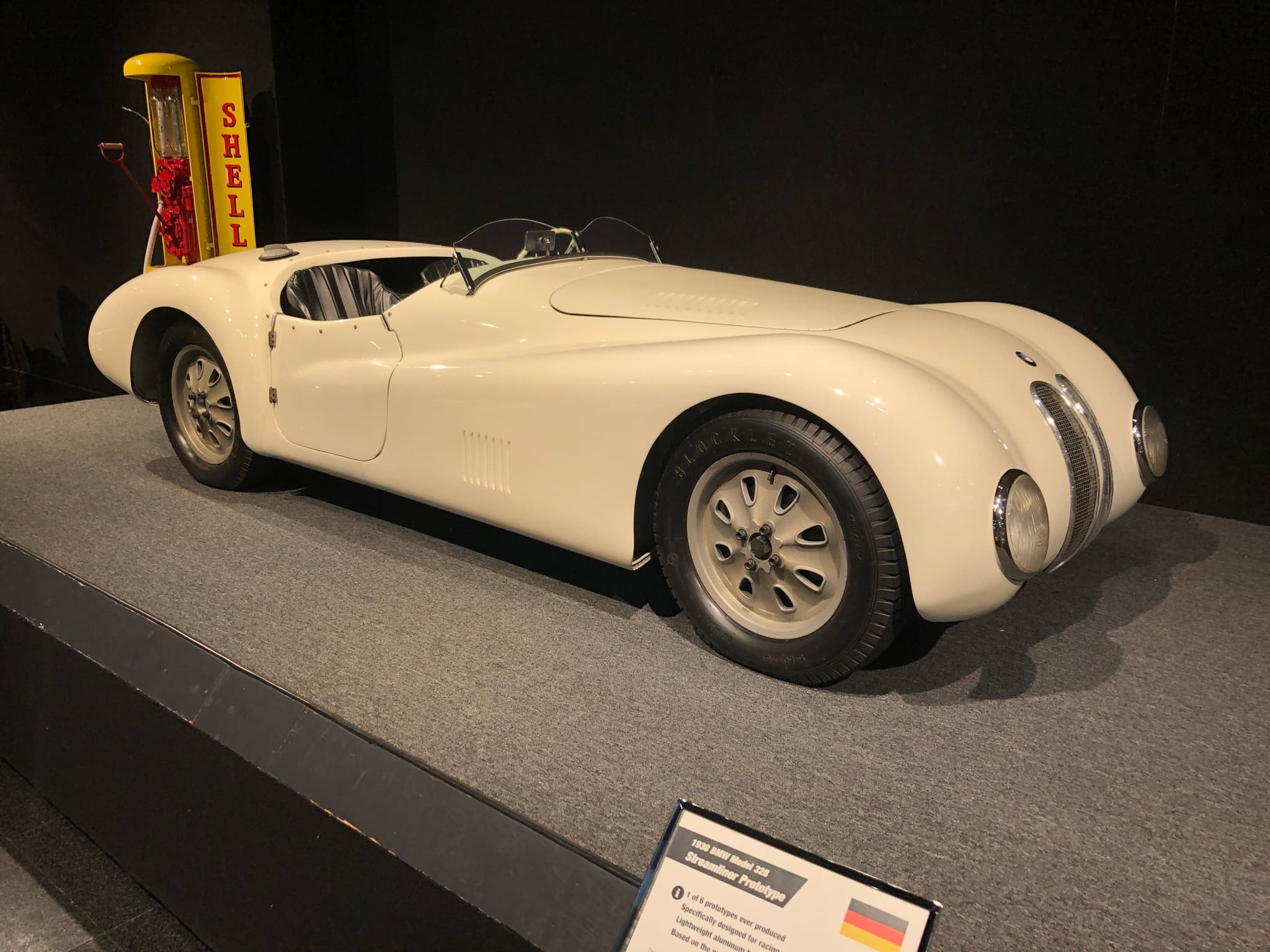 An ivory open streamlined sports car on a museum plinth, a yellow Shell petrol pump behind it.