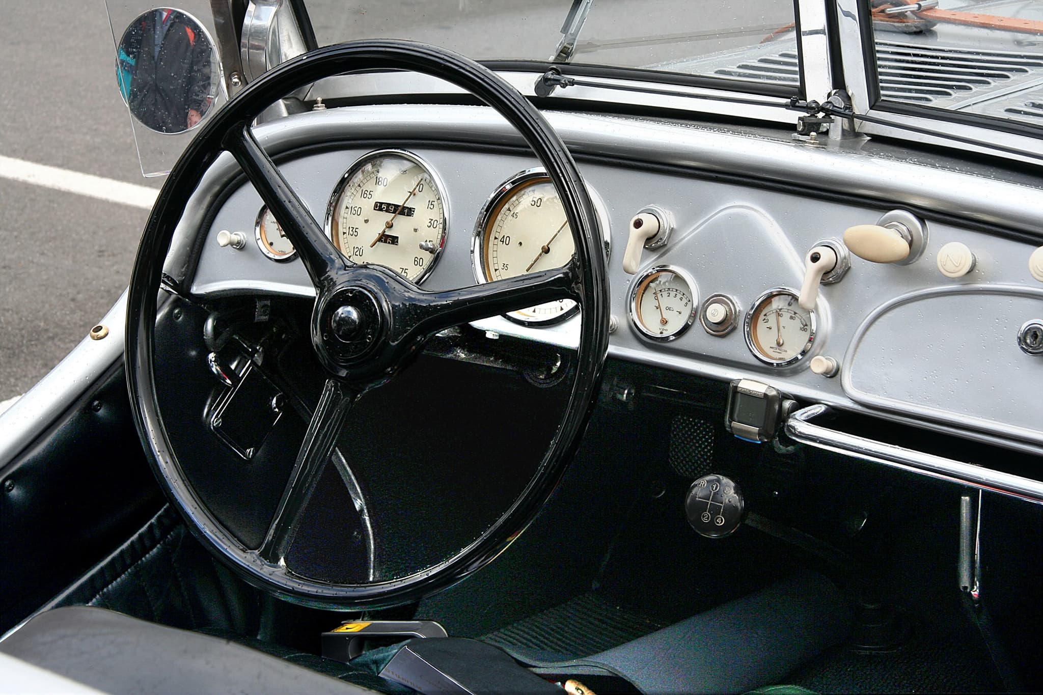 The dashboard of a silver BMW 328 with its three-spoke wheel, two large dials, small gauges and a later trip meter, raindrops on the panel.